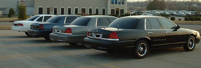 Four sedans parked in a lot, viewed from the rear. The closest is a black 1998 Ford Crown Victoria