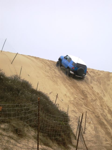 2007 Toyota FJ Cruiser - A blue 2007 Toyota FJ Cruiser with a white camper shell navigates a steep sand dune on an overcast day.