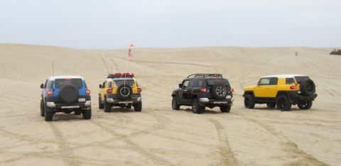 2007 Toyota FJ Cruiser - Four Toyota FJ Cruisers are seen driving on sand dunes under an overcast sky. The vehicles are viewed from the rear.