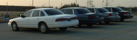 1998 Ford Crown Victoria - A white 1998 Ford Crown Victoria is seen from the side, parked in a row with several other similar vehicles.