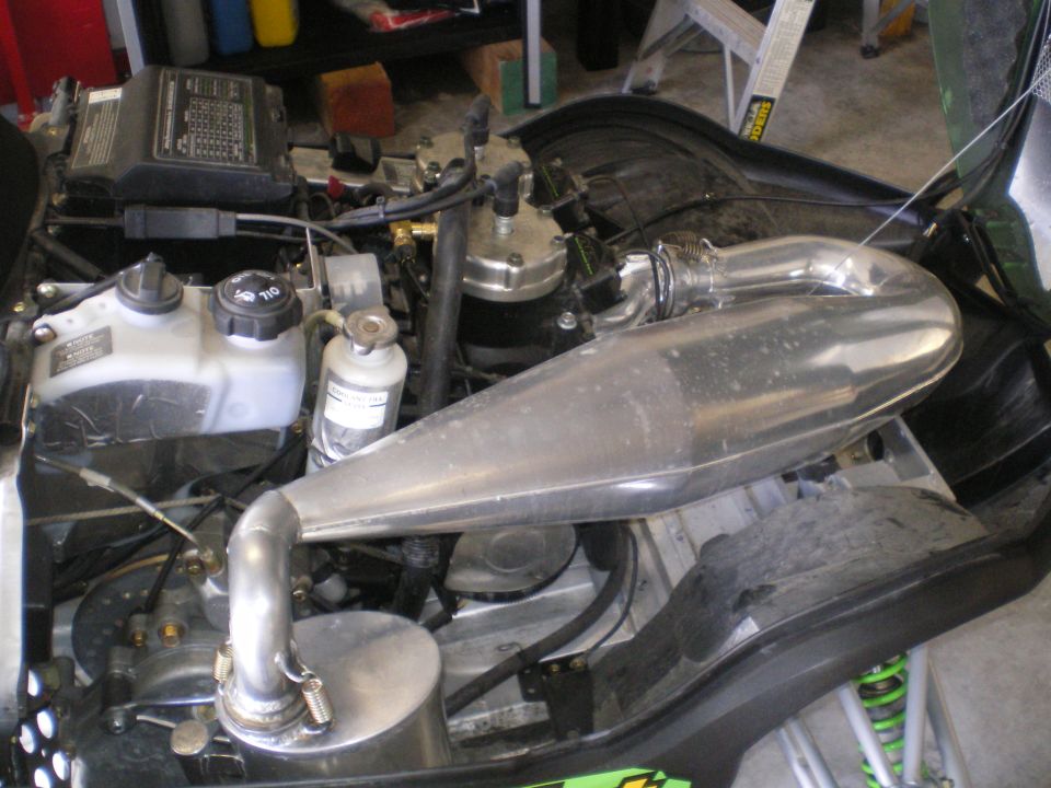 Snowmobile engine bay showing polished aluminum tuned exhaust pipe over two-stroke engine in a garage