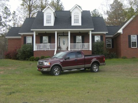 2005 Ford F-150 - A maroon Ford F-150 pickup truck is parked on the lawn in front of a large brick house with a white porch.