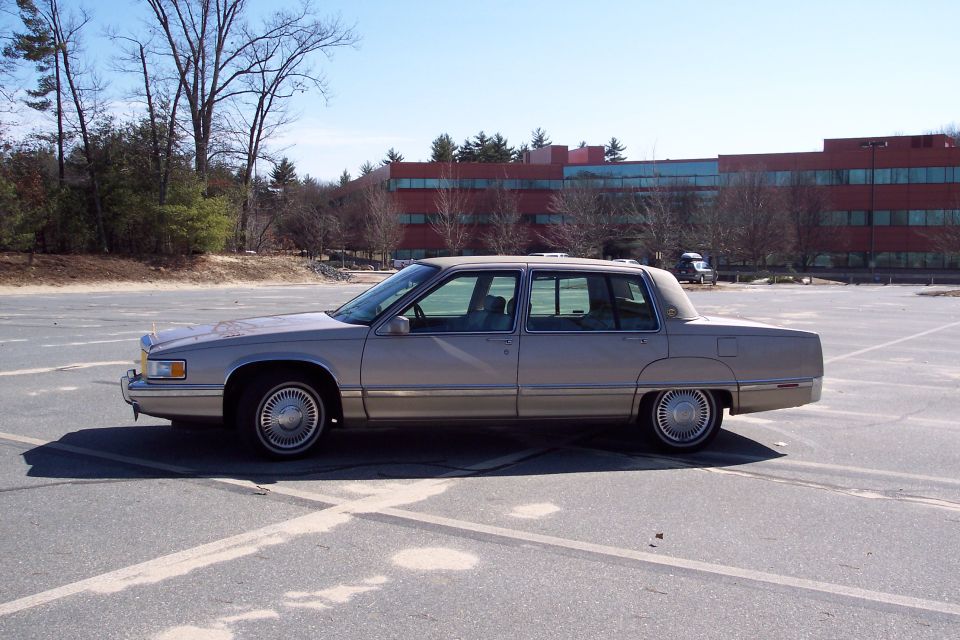 Tan 1993 Cadillac sedan, side view, parked in an empty lot with a red brick building in the background