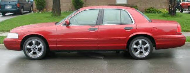 Side view of a red Ford Crown Victoria sedan with chrome five-spoke wheels parked on a wet street