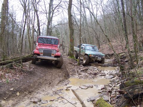 2002 Ford Sport Trac - Two off-road vehicles navigate a challenging, wet path surrounded by trees.