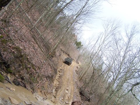 2002 Ford Sport Trac - A dark colored SUV navigates a challenging muddy trail through a wooded area, likely an off-roading excursion.