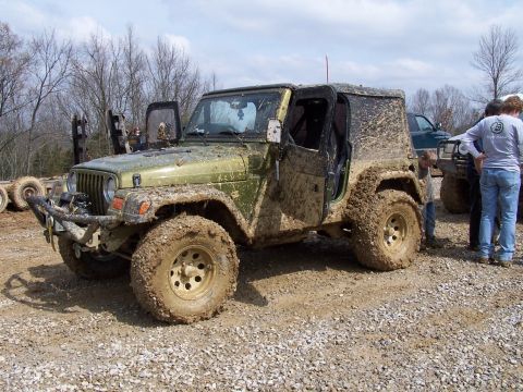 2002 Ford Sport Trac - A mud-covered Jeep Wrangler sits off-road, its doors removed, with other vehicles and people in the background.