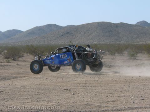 1990 Ford Bronco - blue open-cockpit off-road buggy numbered 1210 kicks up dust racing across flat desert scrub backed by low mountains.
