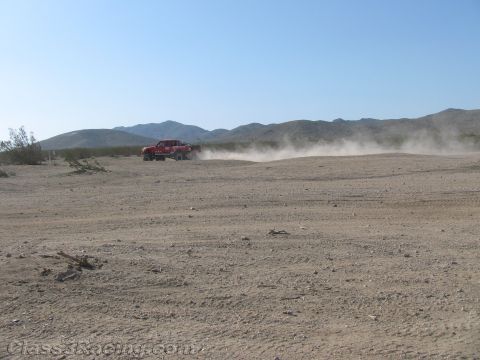 1990 Ford Bronco - red off-road race truck trails a long plume of dust as it speeds across a wide flat desert basin under a clear sky.