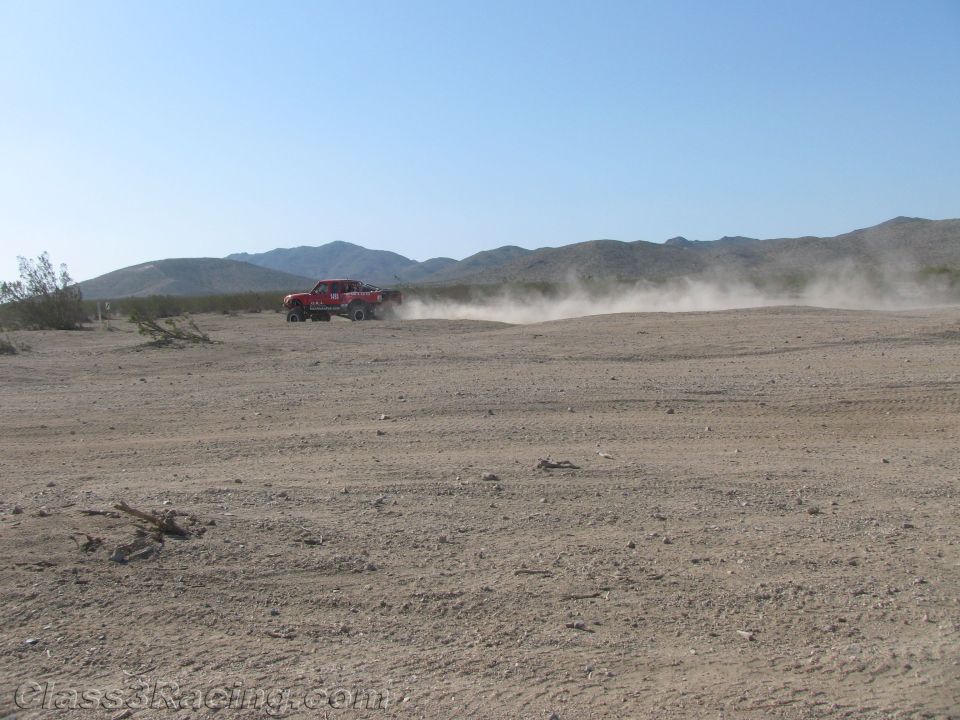 Red off-road truck raising a dust trail in the distance across open desert