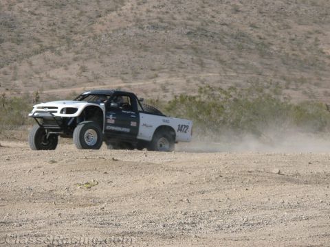 1990 Ford Bronco - white off-road race truck numbered 1472 powers through a dusty turn on a desert course below a barren hillside.