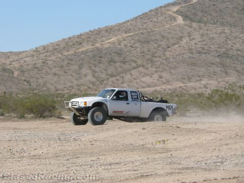 1990 Ford Bronco - white off-road race truck numbered 1464 races over sandy desert terrain with a low mountain rising in the background.