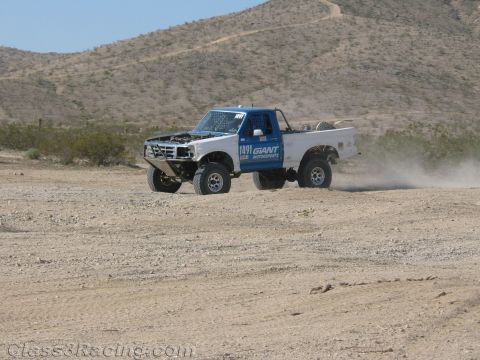 1990 Ford Bronco - white off-road race truck numbered 1491 with Giant Motorsports lettering kicks up dust on a desert course below a hillside.