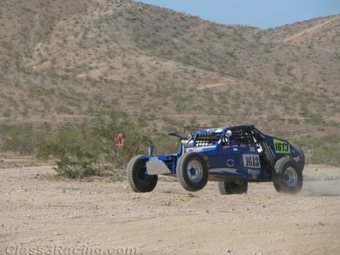 1990 Ford Bronco - blue open-wheel tube-frame off-road buggy numbered 1613 races across a dusty desert course backed by rugged hills.