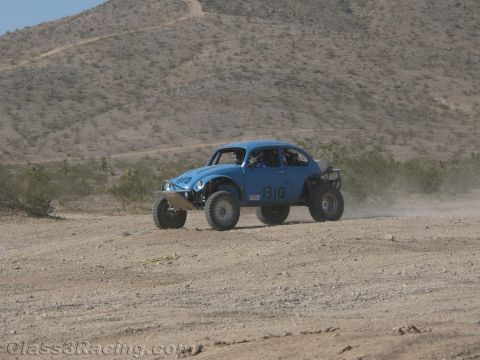 1990 Ford Bronco - blue Baja-style Volkswagen Beetle race car numbered 1310 churns through sandy desert terrain below a low mountain.