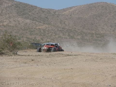 1990 Ford Bronco - Two off-road race vehicles, one orange, run nose to tail through thick dust on a desert course beneath rugged hills.