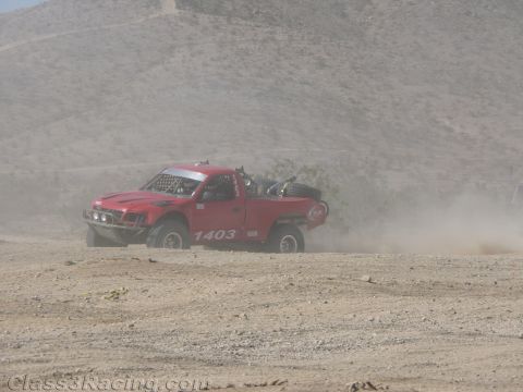 1990 Ford Bronco - red off-road race truck numbered 1403 kicks up a wall of dust as it speeds along a desert course below a barren slope.