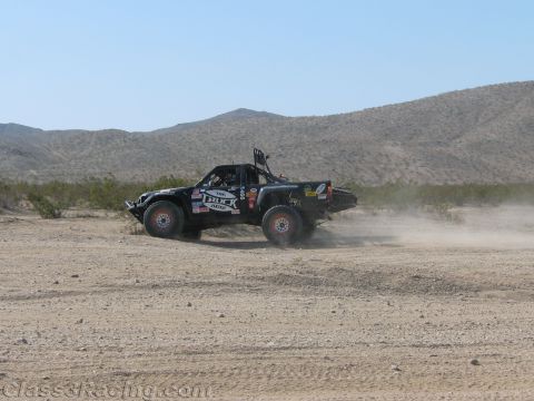 1990 Ford Bronco - black off-road race truck trails dust as it speeds across flat desert scrub backed by distant low mountains.