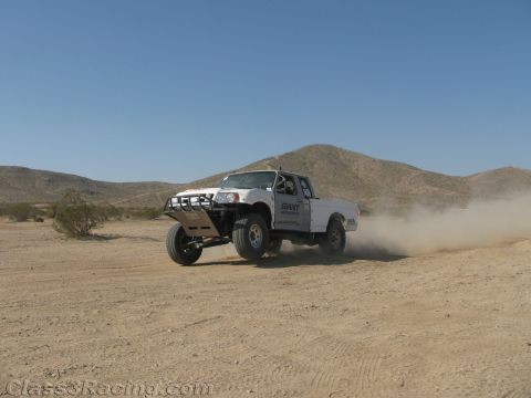 1990 Ford Bronco - white off-road race truck with Giant Motorsports lettering raises dust on a sandy desert course below low mountains.