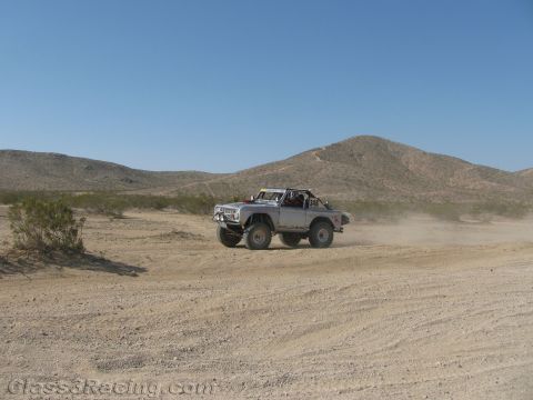 1990 Ford Bronco - silver early-Bronco-style off-road truck races across open desert sand, trailing dust beside a conical hill.