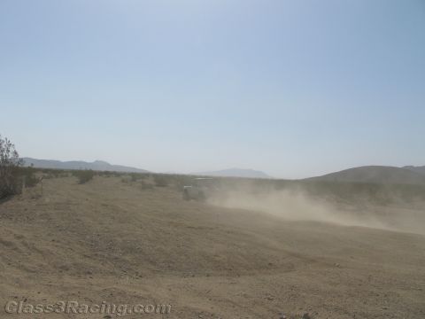 1990 Ford Bronco - off-road race vehicle is almost obscured by a large dust cloud as it crests a sandy berm on a hazy desert course.