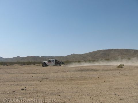 1990 Ford Bronco - white off-road race truck trails a long dust plume as it speeds across a broad flat desert basin under a clear blue sky.