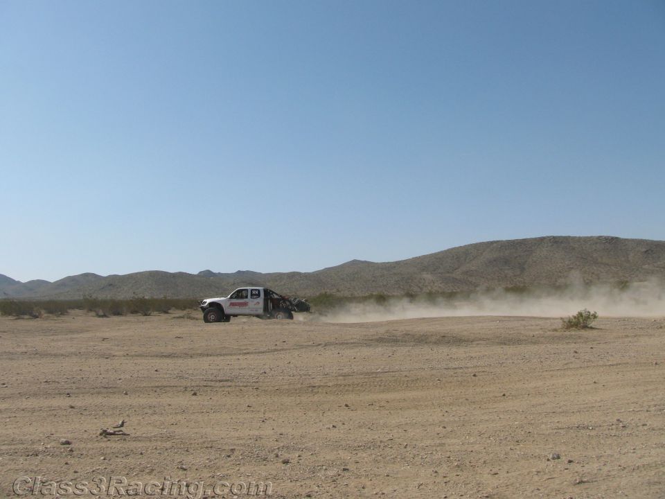 White off-road race truck trailing dust across a wide desert basin