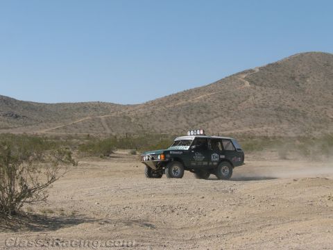 1990 Ford Bronco - teal and white early-Bronco-style off-road race truck with a roof light bar runs a sandy desert course below scrubby hills.