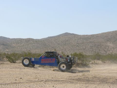 1990 Ford Bronco - blue and red tube-frame off-road race buggy speeds across an open desert flat with dust trailing behind it.