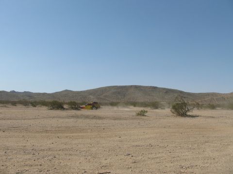 1990 Ford Bronco - small yellow off-road vehicle raises a faint dust cloud while crossing a vast, flat desert plain under a clear blue sky.