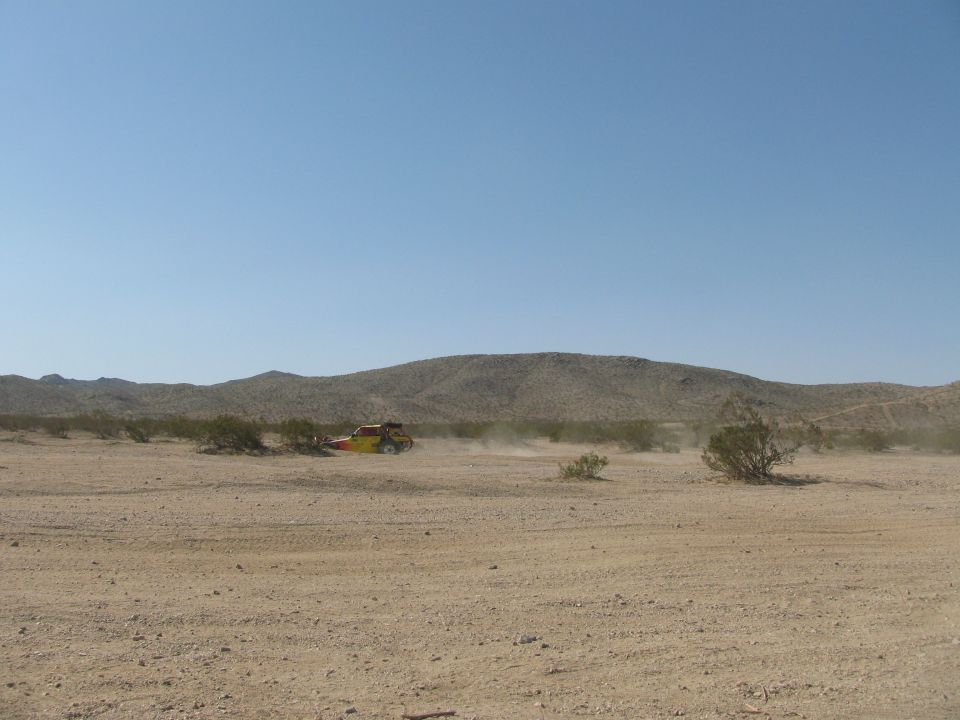 Distant yellow off-road vehicle racing across a wide desert plain