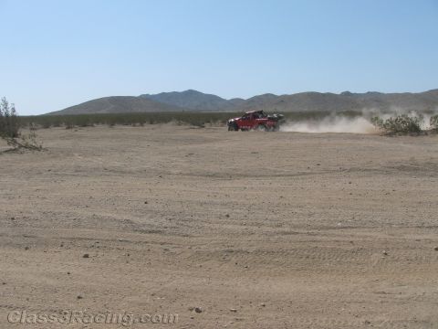 1990 Ford Bronco - red off-road race truck trails dust as it crosses a broad desert flat beneath low mountains.