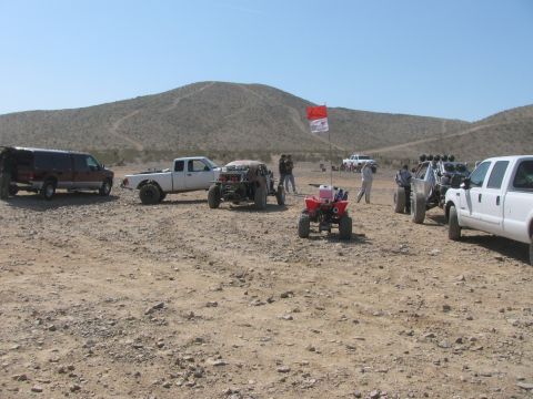1990 Ford Bronco - Off-road race trucks, an ATV, and spectators gather at a desert staging area marked by a red flag near low hills.
