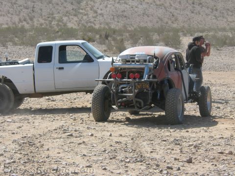 1990 Ford Bronco - red Baja-style Volkswagen Beetle off-road car with exposed engine sits beside a white pickup truck on rocky desert ground.