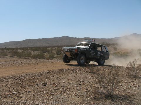 1990 Ford Bronco - black SUV-bodied off-road race truck catches air on a rocky desert trail with a plume of dust behind it.
