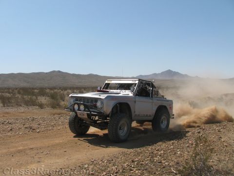 1990 Ford Bronco - silver early Ford Bronco off-road racer powers through desert dirt, kicking up a large spray of dust.