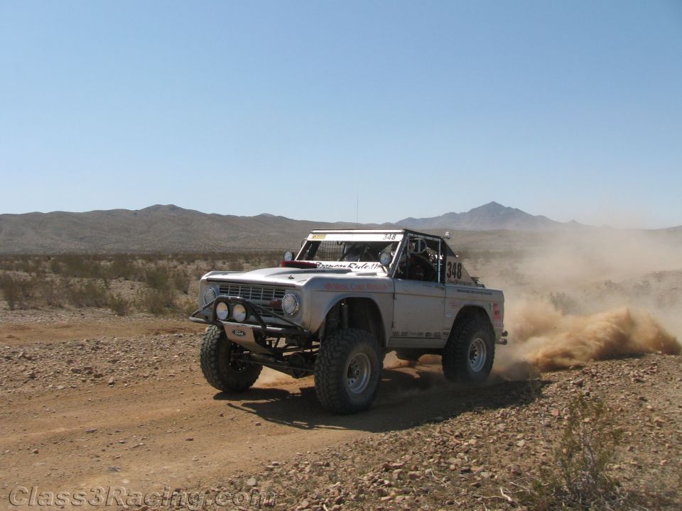 Silver early Ford Bronco off-road racer powering through desert dirt