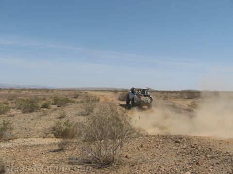 1990 Ford Bronco - off-road race buggy charges head-on across brushy desert terrain, trailing a wide cloud of dust.