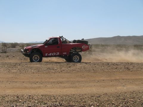 1990 Ford Bronco - red off-road race truck marked 1403 speeds along a dirt desert road with dust rising behind it.