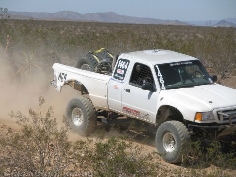 1990 Ford Bronco - white off-road race truck numbered 1464 leaps through dense desert brush with its front wheels lifted.