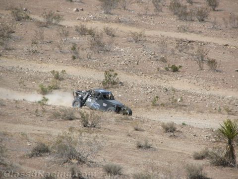 1990 Ford Bronco - gray off-road race car travels along a winding hillside desert trail dotted with scrub and rocks.