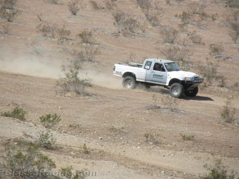 1990 Ford Bronco - white off-road race truck speeds down a sloped, scrub-covered desert trail, raising a long dust trail.