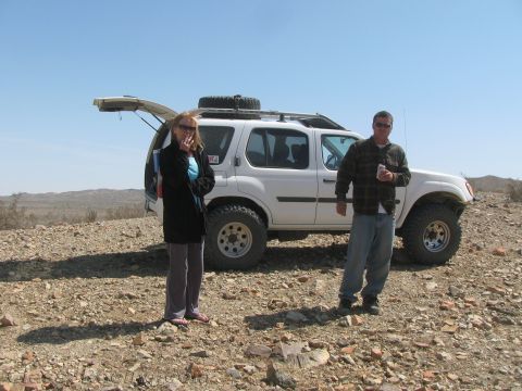 1990 Ford Bronco - woman and a man stand on rocky ground beside a white SUV fitted with a rooftop spare tire and roof rack.