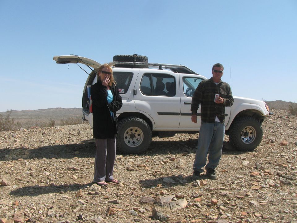 Two people standing by a white SUV with rooftop spare tire in the desert