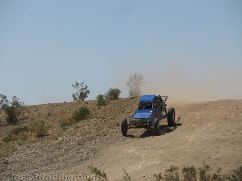 1990 Ford Bronco - blue tube-frame off-road race buggy descends a bare dirt hillside, trailing a faint cloud of dust.