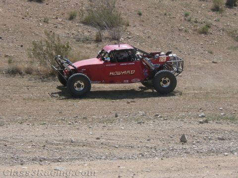 1990 Ford Bronco - red tube-frame off-road race buggy lettered HOWARD drives across hard-packed desert ground below a slope.