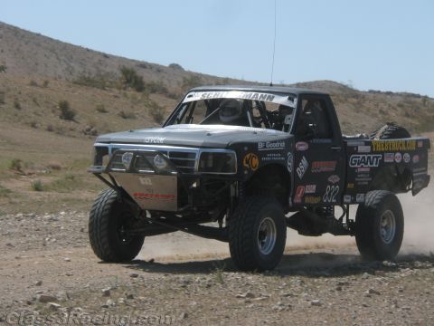 1990 Ford Bronco - black off-road race truck numbered 832 and covered in sponsor decals drives across a desert course near hills.