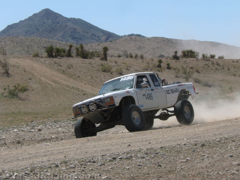 1990 Ford Bronco - white off-road race truck numbered 1486 races along a dirt course with a rocky mountain peak in the background.