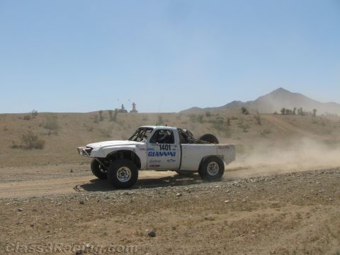 1990 Ford Bronco - white off-road race truck marked 1401 with GIANNINI lettering travels a dirt road near a desert mountain peak.