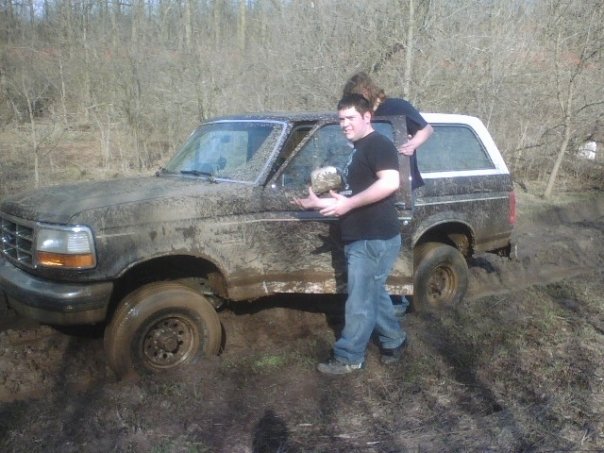 Mud-covered black Ford Bronco with white top in muddy field, two young men at driver's side
